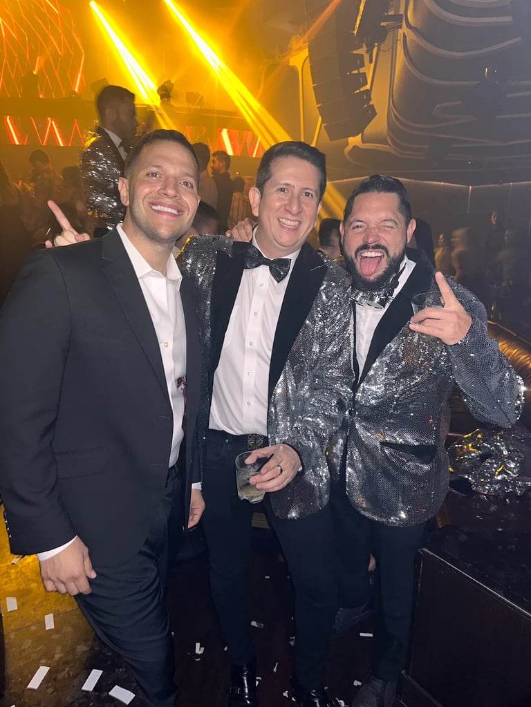 Three men in formal attire at a nightclub with gold neon lighting in the background, posing together and smiling for the camera